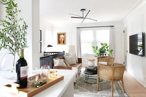 A modern living room with a sofa, two chairs, a coffee table, and a large window, featuring plants and artwork.