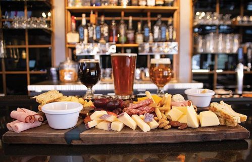A wooden platter with assorted cheeses, meats, nuts, and grapes, accompanied by two glasses of beer in a bar setting.
