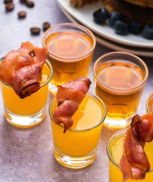 A close-up of shot glasses filled with orange liquid, each topped with a piece of crispy bacon.