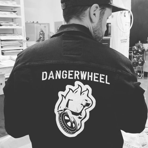 A person wearing a black jacket with "DANGERWHEEL" and a graphic design on the back, standing in an artistic space.