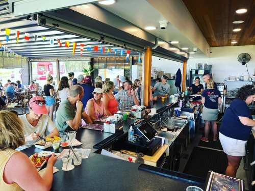 A busy bar scene with patrons enjoying food and drinks, colorful decorations, and staff serving at the counter.