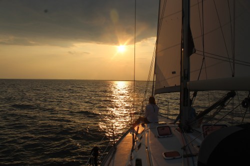 A person sits on a sailboat, watching the sunset over calm waters, with golden reflections on the surface.