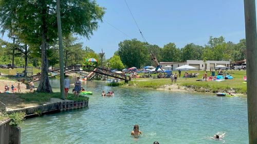 A sunny day at a water park with people swimming, lounging, and a zip line over a clear blue water area.