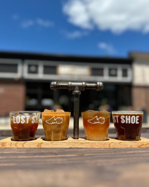 Four craft beer samples in glasses on a wooden flight board, with a sunny outdoor backdrop and blue sky.