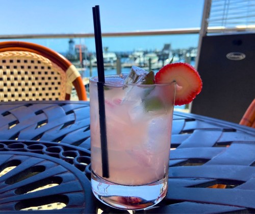 A refreshing cocktail with ice, garnished with a strawberry and lime, sits on a table overlooking a marina.