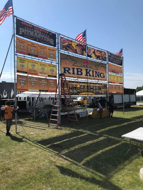A large sign for "Carolina Rib King" at a barbecue event, featuring awards and flags, with people setting up nearby.