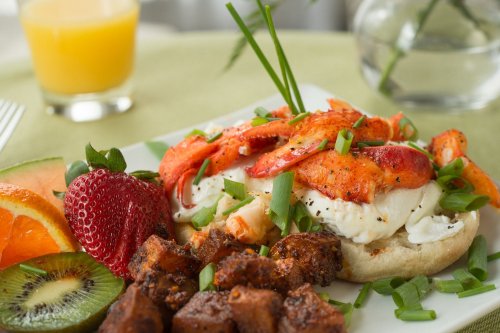 A plate of poached eggs topped with lobster, served with fruit and seasoned potatoes, alongside a glass of juice.