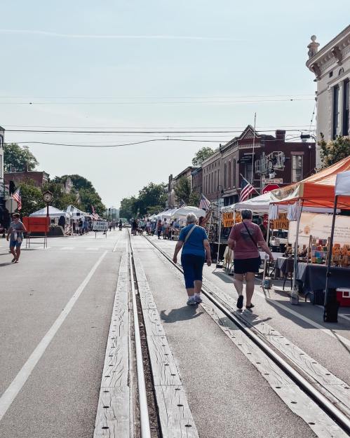 Two women walk along a street lined with vendor tents and American flags, with train tracks running through the center.