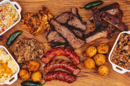 A wooden platter featuring various meats, sides, and jalapeños, including brisket, ribs, sausage, and cornbread balls.