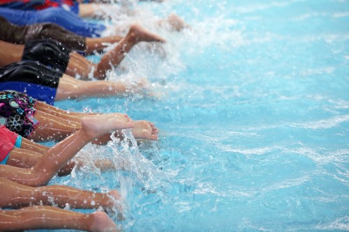 A close-up of multiple feet splashing in a swimming pool, creating waves in the clear blue water.