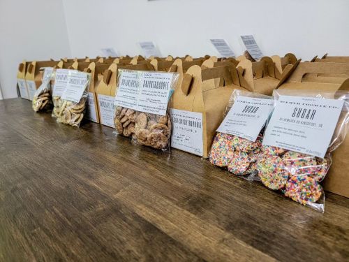 A wooden table displays neatly packaged snacks in brown boxes and clear bags, labeled with various flavors.