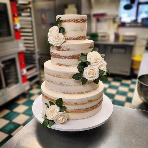 A three-tiered wedding cake with white frosting, gold accents, and white roses, set in a kitchen.
