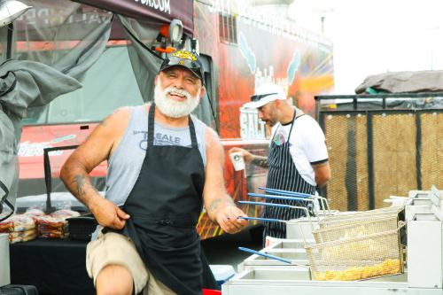A smiling chef in an apron cooks at a food truck, with another chef in the background preparing food.