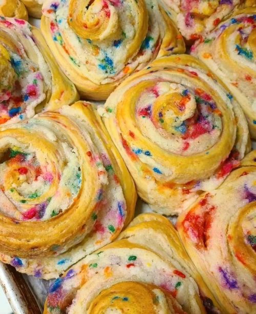Colorful, swirled cinnamon rolls with vibrant sprinkles, arranged closely on a baking tray.