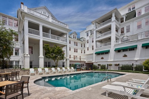 A serene pool area surrounded by white buildings, with lounge chairs and greenery under a blue sky.