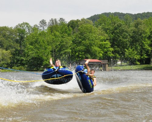 Two people are enjoying tubing on a lake, splashing water as they ride on inflatable tubes. Green trees surround the area.