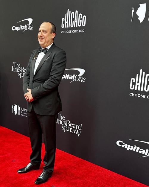 A man in a tuxedo smiles on a red carpet at an event with a black backdrop featuring logos and text.