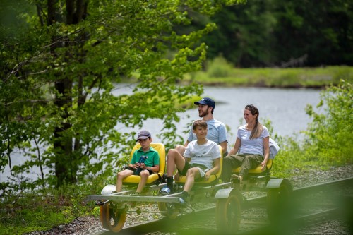 A family enjoys a ride on a pedal-powered rail cart along a scenic track by a lake, surrounded by greenery.