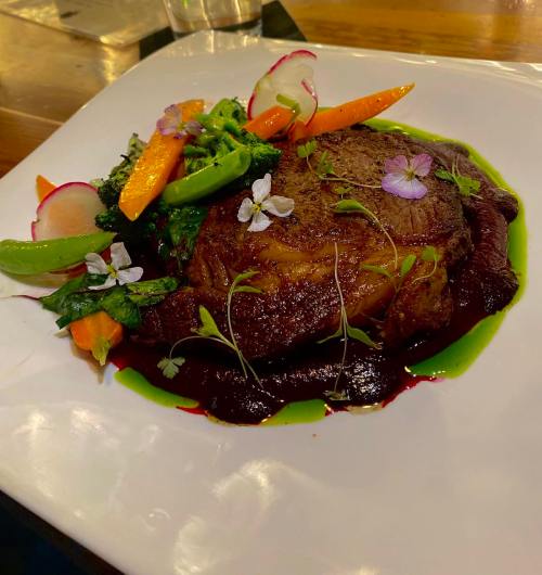 A beautifully plated steak with colorful vegetables and edible flowers on a white dish.