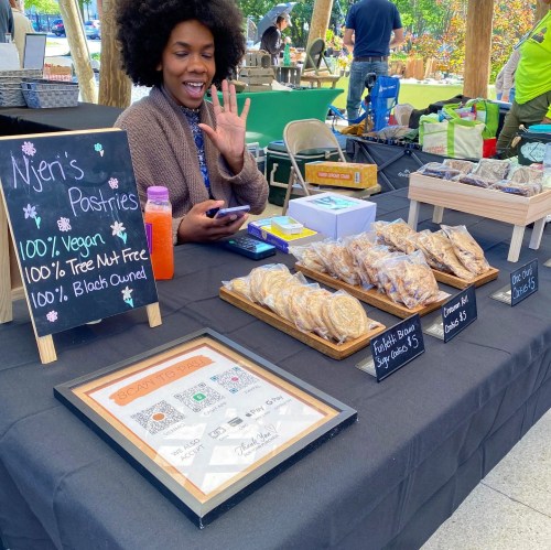 A smiling vendor waves from behind a table displaying vegan pastries and a sign for Njen's Pastries at a market.