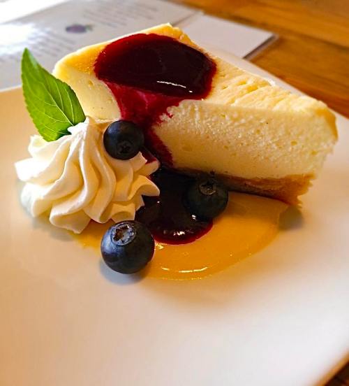 A slice of cheesecake topped with berry sauce, fresh blueberries, and a dollop of whipped cream on a white plate.
