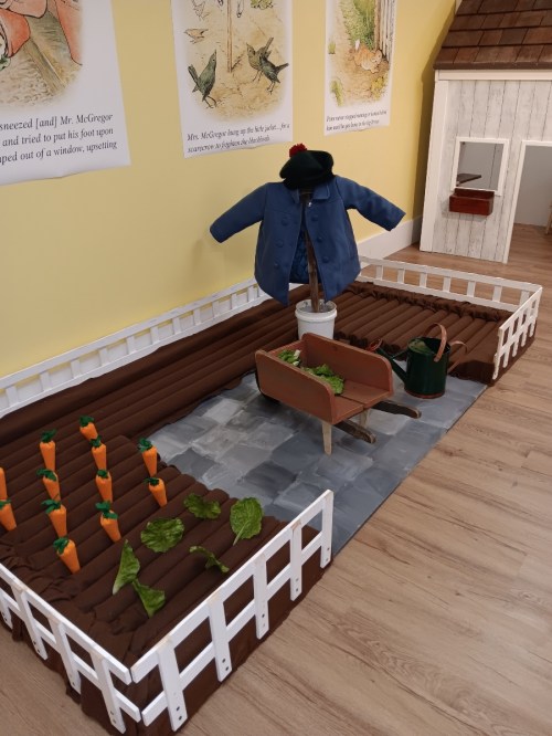 A miniature garden scene with a blue coat, wheelbarrow, watering can, and vegetable patches of carrots and lettuce.