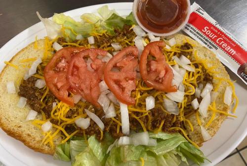 A plate with a large sandwich topped with chili, lettuce, cheese, tomatoes, onions, and a side of sour cream and sauce.