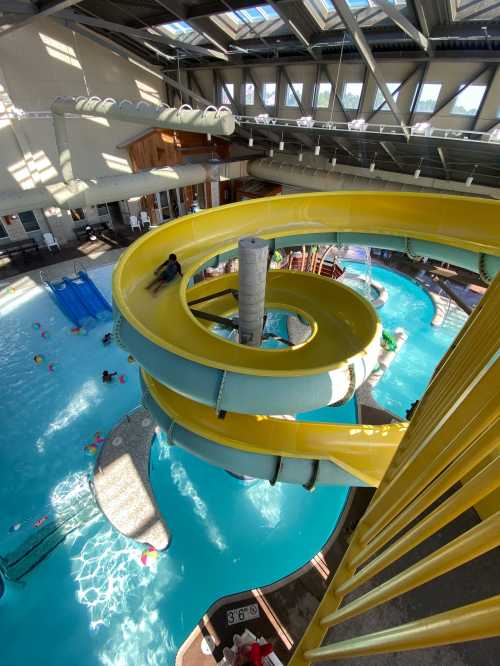 A colorful water slide spirals into a pool area with children playing and floating toys in a bright indoor setting.