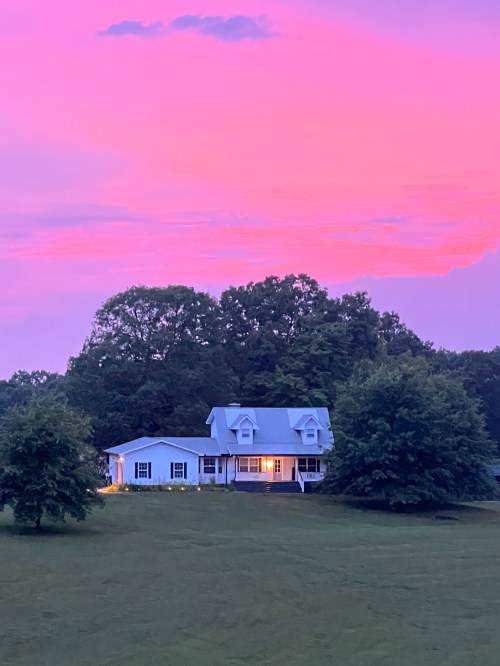 A charming white house with a porch, surrounded by trees, under a vibrant pink and purple sunset sky.