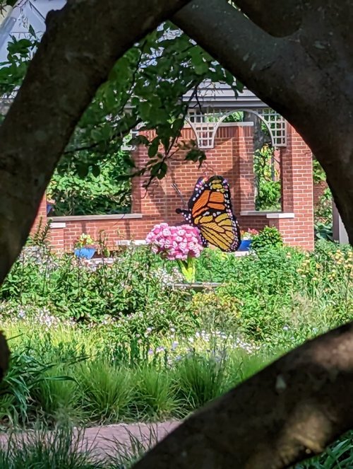 A vibrant butterfly sculpture near pink flowers, framed by tree branches in a lush garden setting.