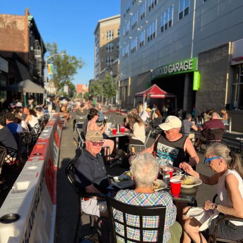 A group of people dining outdoors at tables on a sunny street, with buildings and trees in the background.