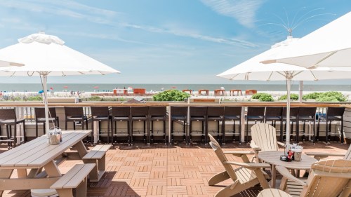 A sunny beachside patio with umbrellas, tables, and a view of the ocean and beach chairs in the distance.