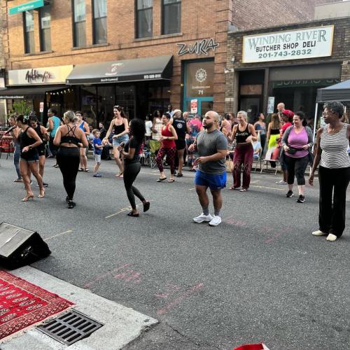 A diverse group of people dancing in the street during a lively outdoor event, with shops in the background.