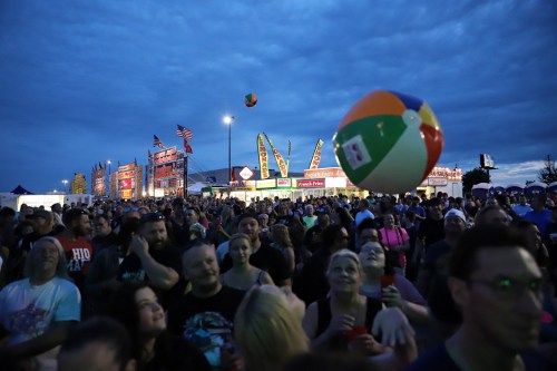 A crowd enjoying a festival at dusk, with colorful booths and a beach ball in the air.