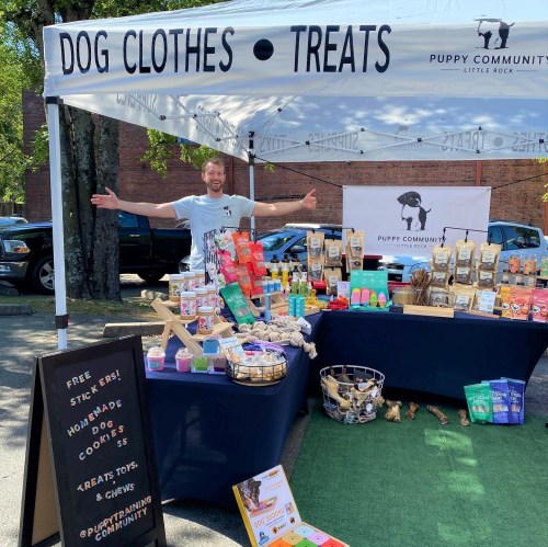 A vendor at a market showcases dog clothes and treats under a tent, with a sign for Puppy Community in Little Rock.