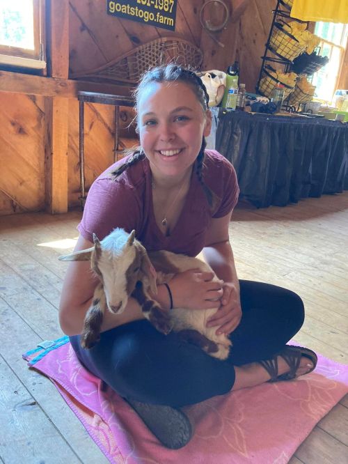 A smiling person sits on a mat, holding a small goat in a cozy indoor setting with wooden walls.