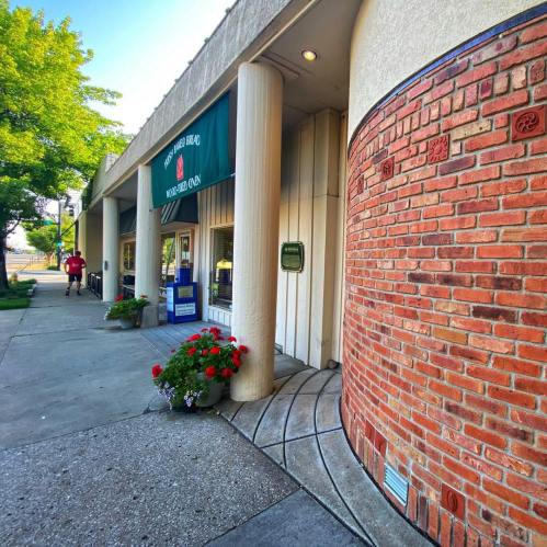A storefront with a green awning, brick accents, and flower pots, featuring a sidewalk and a person walking by.