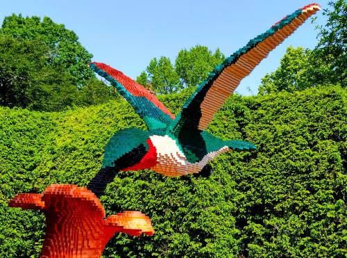 A colorful bird sculpture made of LEGO bricks, hovering above a flower in a lush green garden.