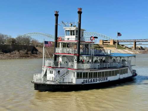 A large riverboat named "Island Queen" sails on a muddy river, with flags flying and a bridge in the background.