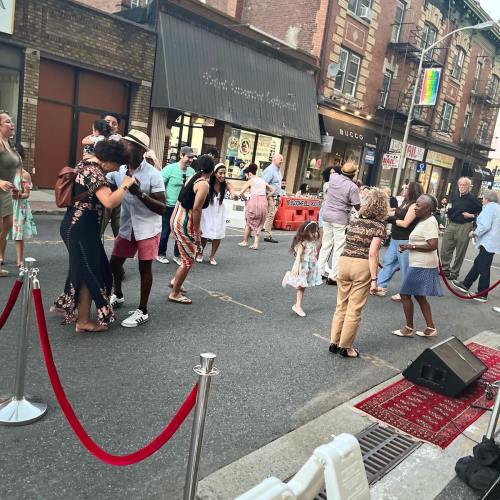 A lively street scene with people dancing and socializing, surrounded by shops and festive decorations.