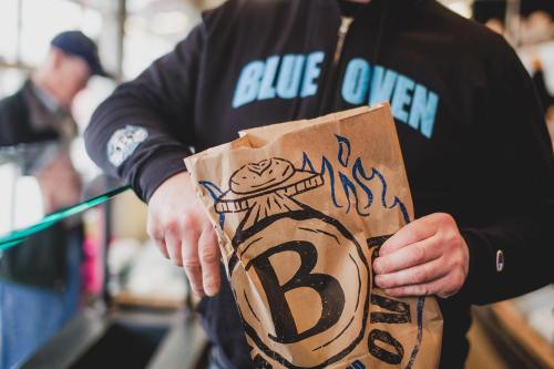 A person in a black hoodie holds a brown paper bag with a logo, inside a bakery or café setting.