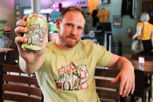 A man in a light green shirt holds a can of pickle-flavored drink, smiling in a casual setting.