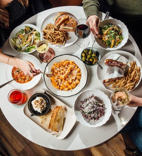 A table filled with various dishes, including pasta, salads, fries, and drinks, with hands reaching for the food.