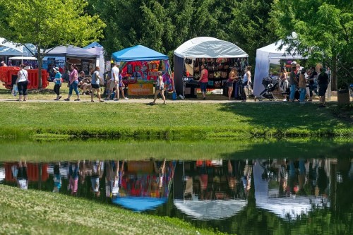 A vibrant outdoor market with colorful tents, people walking, and reflections in a calm pond. Green trees in the background.