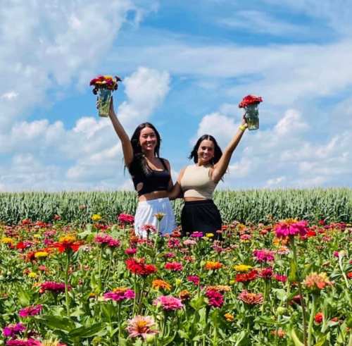 Two women stand in a colorful flower field, each holding a vase of flowers, with a blue sky and clouds above.
