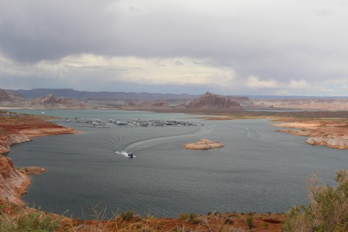 A scenic view of a lake surrounded by rocky cliffs and distant mountains, with a boat on the water under a cloudy sky.