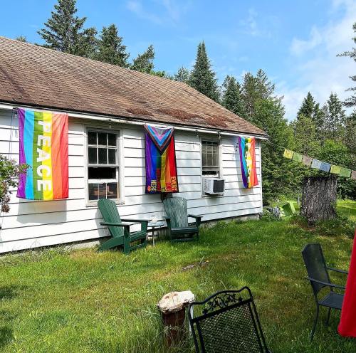 A white house with rainbow flags and a peace flag hanging, surrounded by green grass and trees on a sunny day.