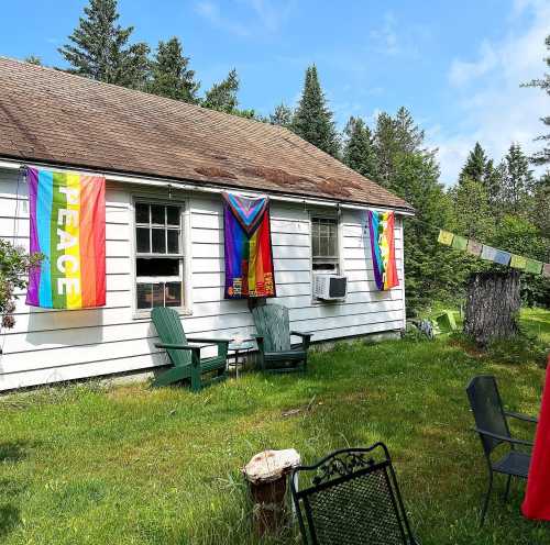 A white house with rainbow flags and a peace flag hanging, surrounded by green grass and trees on a sunny day.