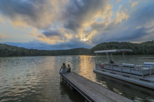 Two people sit on a dock by a calm lake, watching a colorful sunset with clouds overhead and a boat nearby.