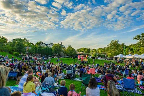 A large crowd enjoys an outdoor event on a grassy area, with tents and a stage under a blue sky with clouds.